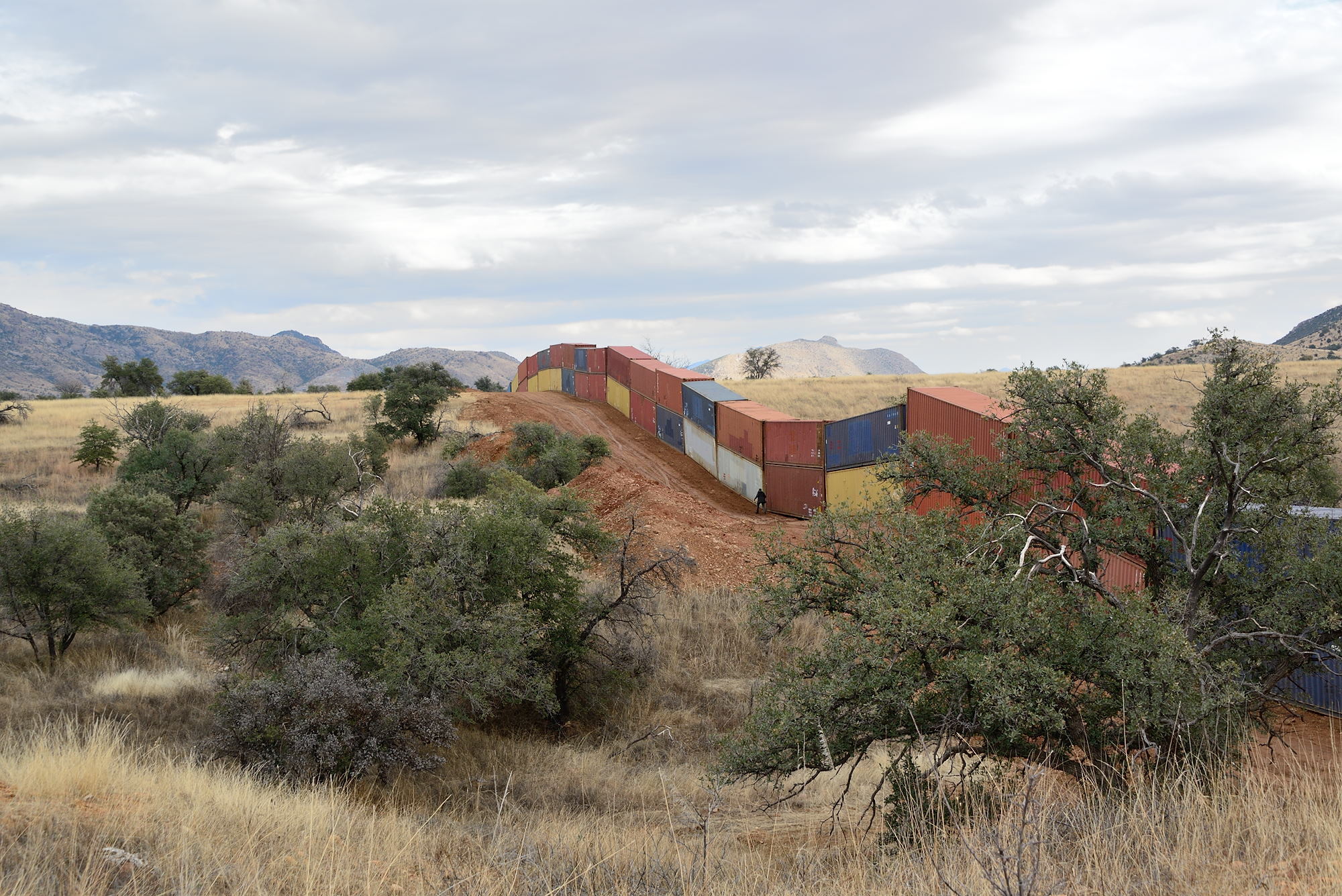 A photograph of a wall of multi-colored shipping containers, two containers high, snakes up and down a hill. There are mountains in the background.
