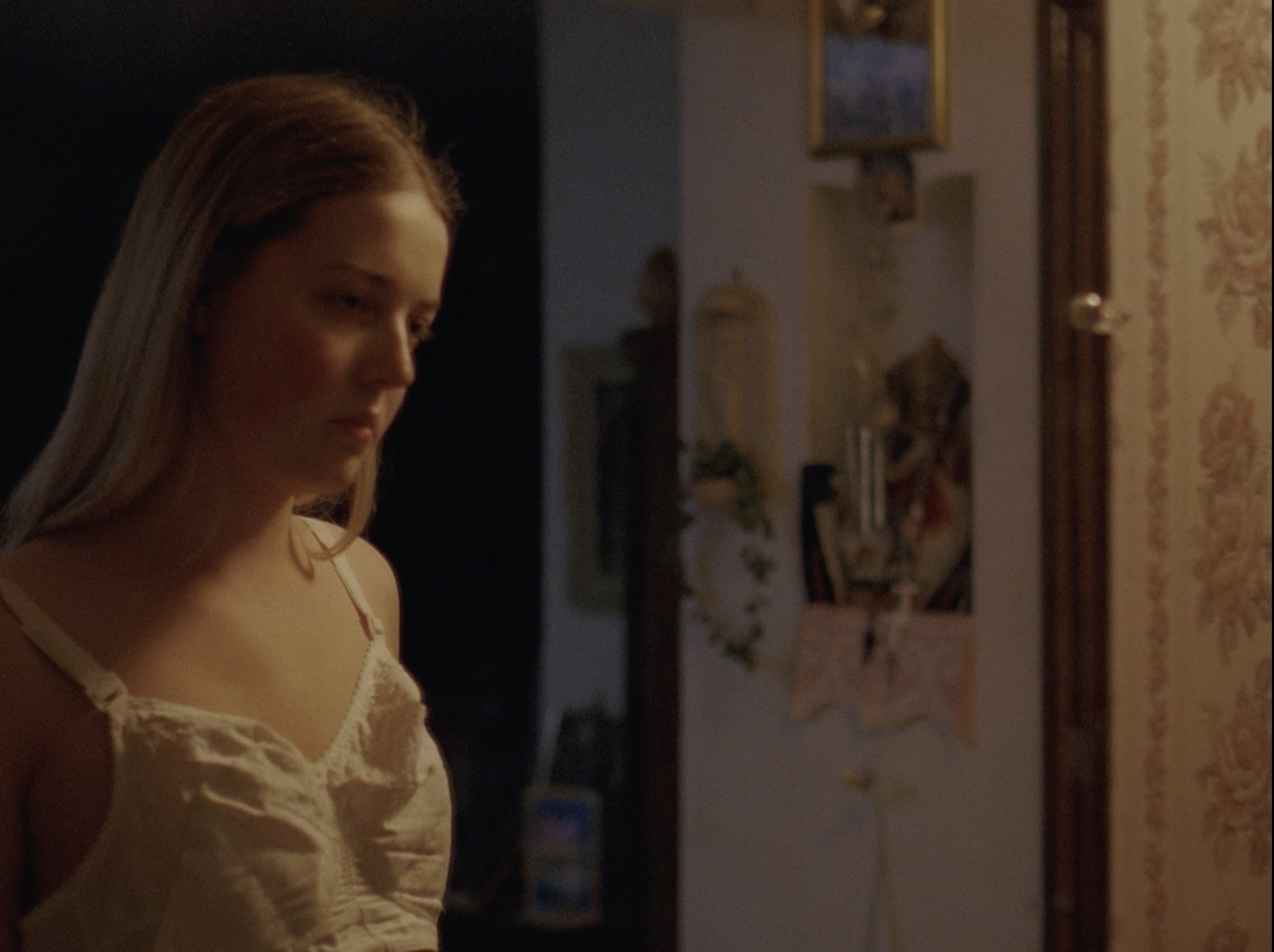 In a darkened hallway, a young white trans woman with long brown hair looks in the mirror at the old-fashioned cotton bra she wears that is too large for her.